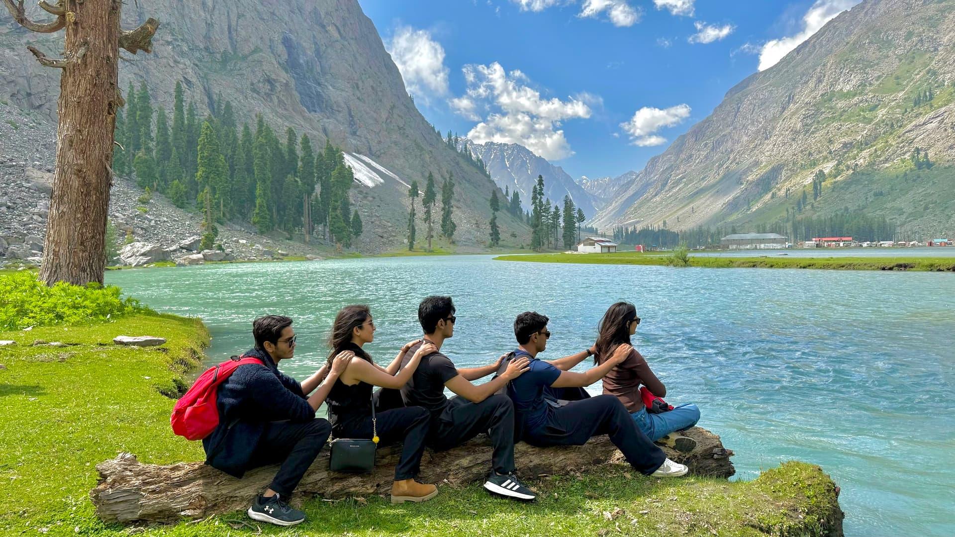 Five travelers sitting together beside a turquoise mountain lake with mountains in the background.